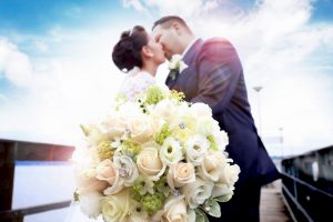 Brautpaar küsst sich auf einem Bootssteg und hält einen Brautstraußin die Kamera